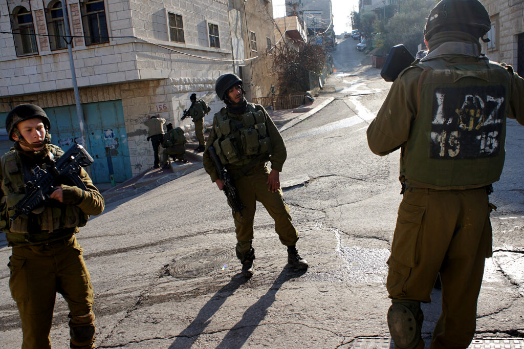 Near a checkpoint in Hebron, Israeli soldiers stand by as their colleagues search a Palestinian man in the background (Friends123/Wikimedia Commons)