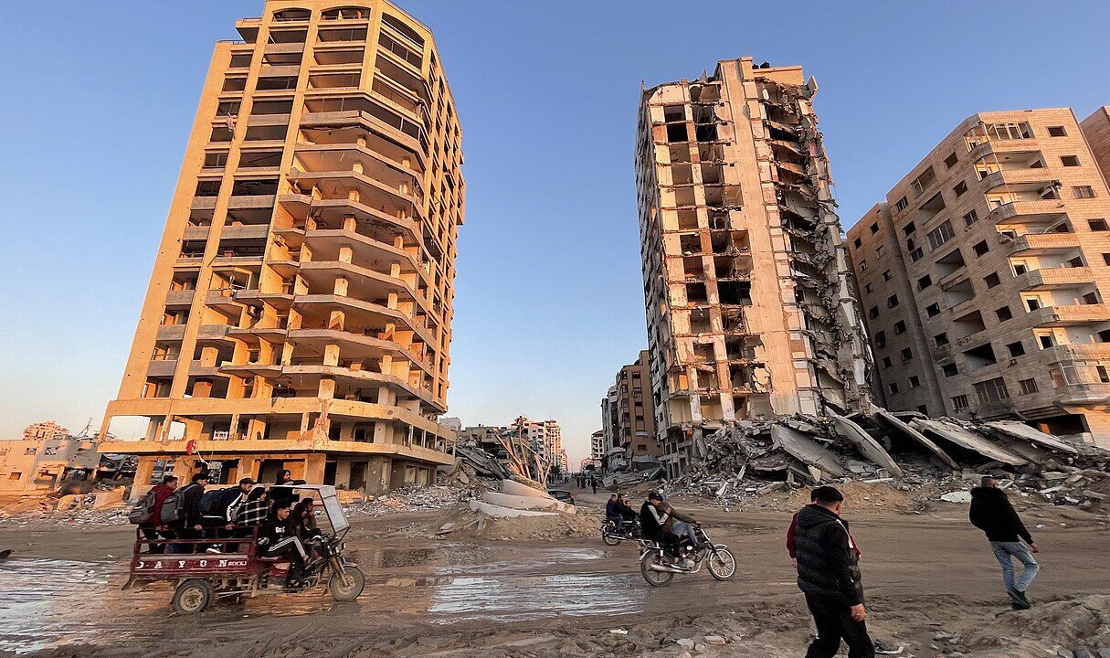 An image from Feb. 14, 2025, shows Palestinians in the Gaza Strip passing bombed-out buildings (Jaber Jehad Badwan/Wikimedia Commons)