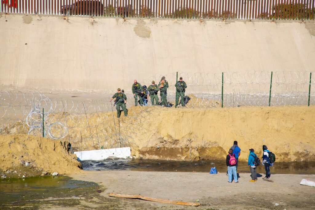 Border Patrol agents tend to concertina wire near the US-Mexico boundary in Tijuana as migrants look on (Humberto Chávez/Unsplash)