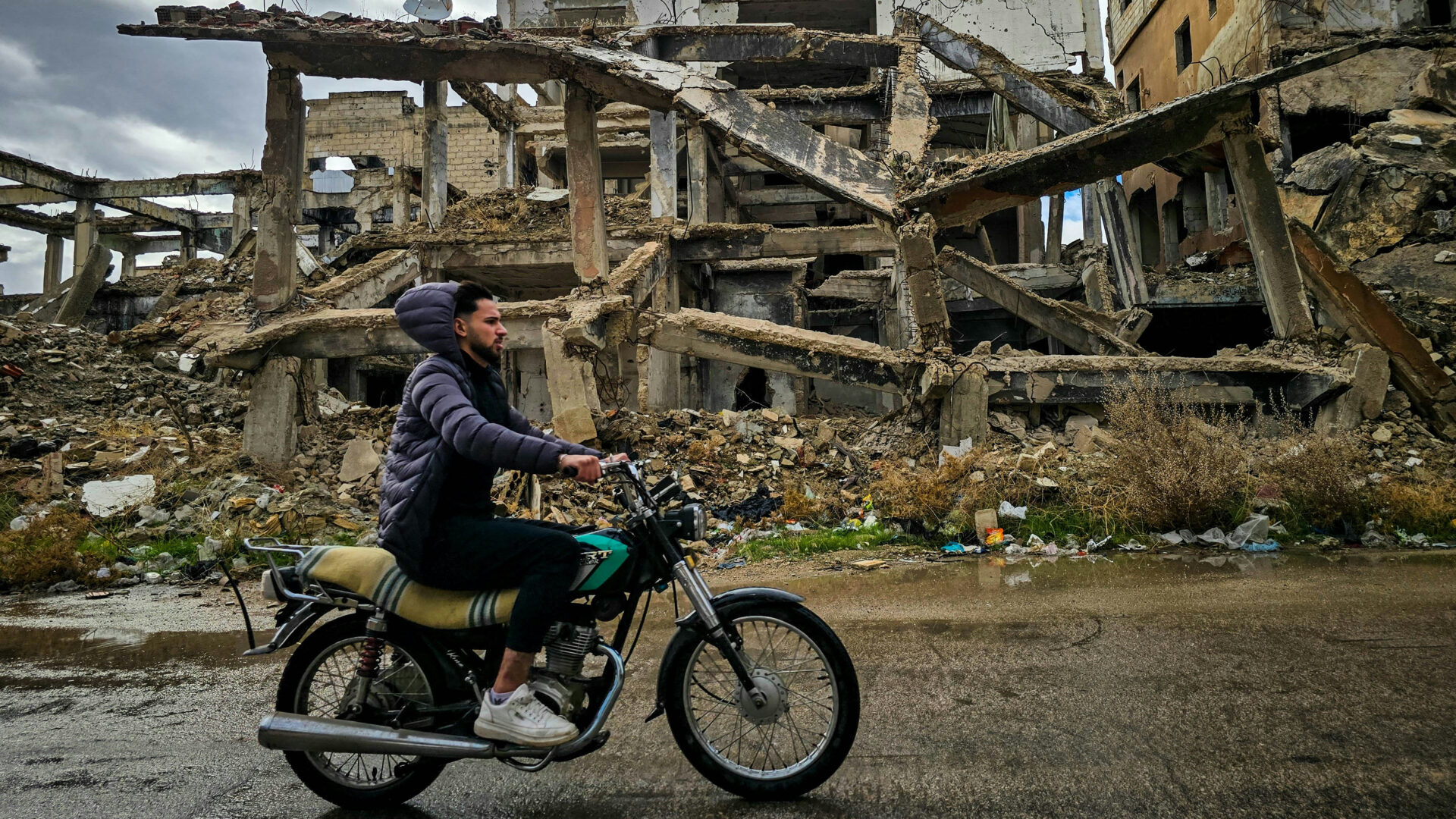 A Syrian man rides his motorbike past a destroyed building in Daraa in February 2025 (Mahmoud Sulaiman/Unsplash)